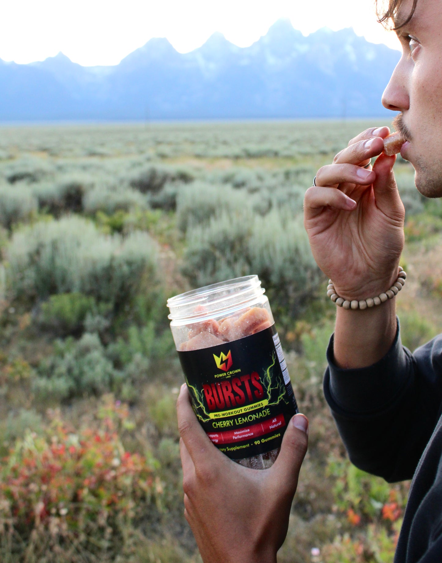 Person eating pre-workout gummies from a container labeled 'Bursts' in a scenic outdoor setting.