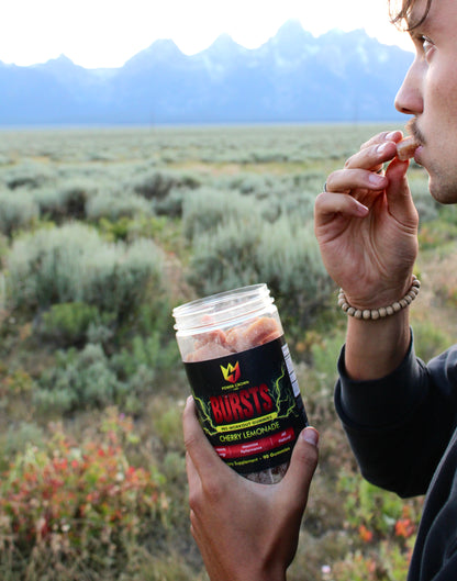 Person eating pre-workout gummies from a container labeled 'Bursts' in a scenic outdoor setting.